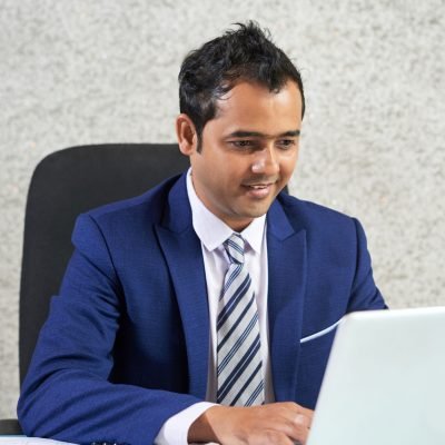 Smiling manager in blue suit sitting at his workplace and typing on laptop at office