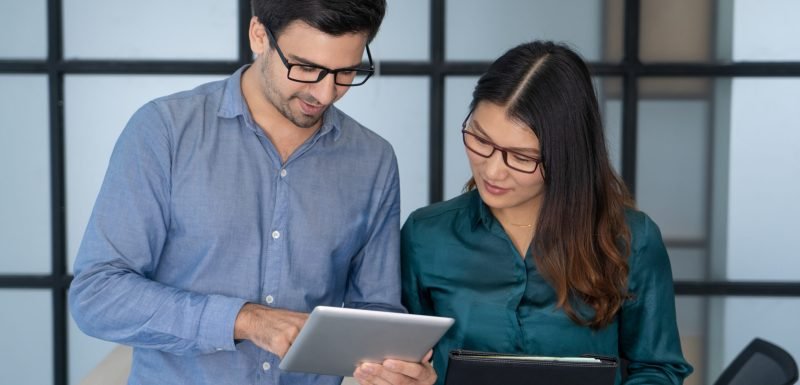 Caucasian supervisor helping new Asian employee. Friendly office worker showing to young colleague project details on tablet screen. Training and corporate help concept