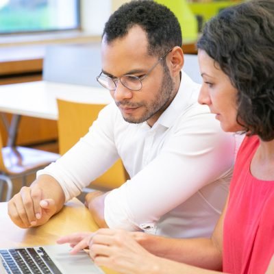 Coworkers doing research in library. Man and woman in casual sitting at desk, using laptop and talking. Teamwork or collaboration concept