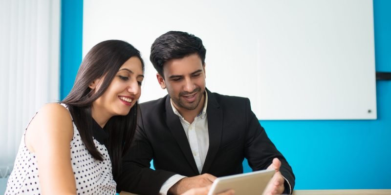 Portrait of happy young colleagues using digital tablet. Young Caucasian businessman and businesswoman sitting at table and watching data on pc tablet. Wireless technology in business