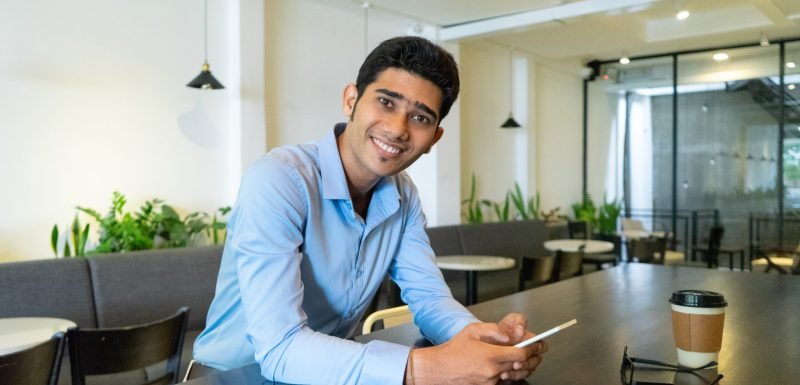 Portrait of happy young Indian businessman sitting in cafe. Young man sitting at table, using mobile phone, looking at camera and smiling indoors. Coffee break or freelance working concept