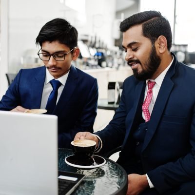 Two indian business man in suits sitting at office on cafe, looking at laptop and drinking coffee.